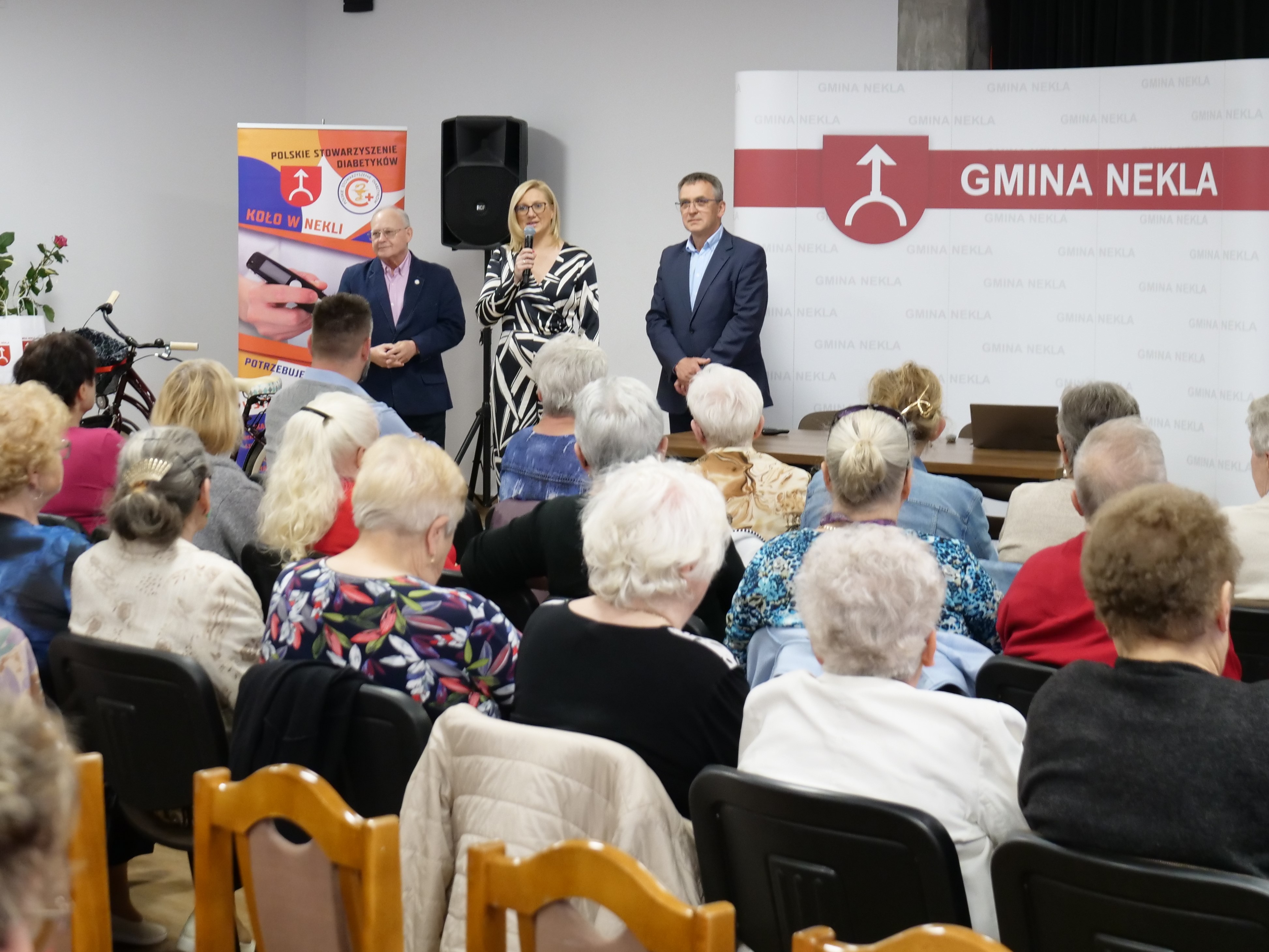 Zdjęcie przedstawia 3 osoby, stojące przed grupą siedzących na krzesłach. Dwóch mężczyzn, pomiędzy nimi kobieta z mikrofonem. W tle baner z napisem GMINA NEKLA oraz drugi z napisem Polskie Stowarzyszenie Diabetyków Koło w Nekli.